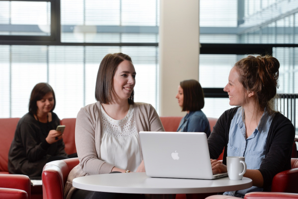 Zwei Frauen sitzen im Hintergrund auf einem Sofa, im Vordergrund sitzen zwei Frauen zu einer Besprechung an einem runden Tisch, auf dem ein aufgeklappter Rechner steht. 