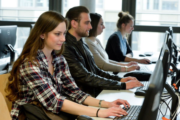 Vier Personen sitzen an einer Tischreihe vor Bildschirmen und arbeiten