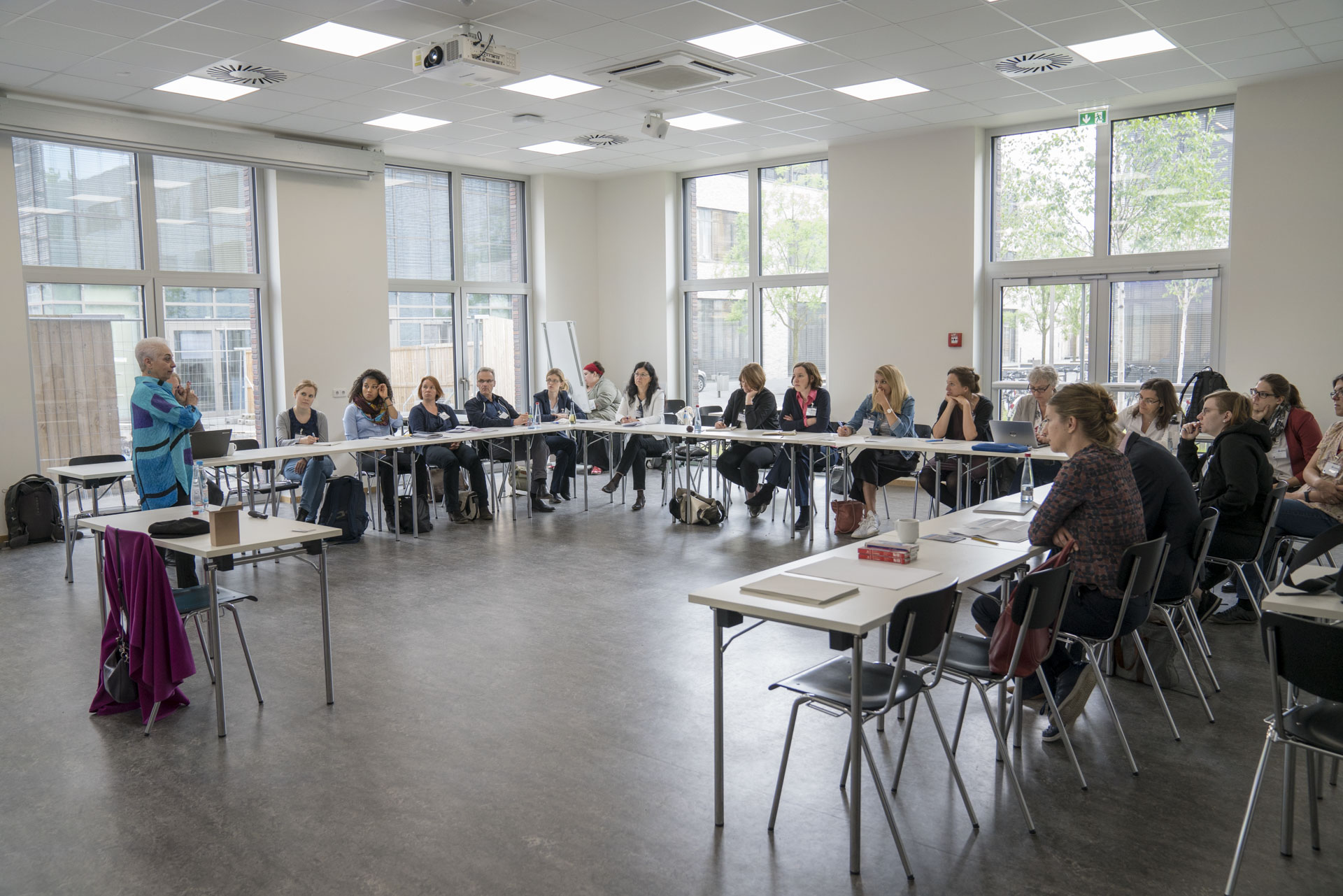 Im Workshop 1.1 Personen sitzen am u-förmig aufgebauten Tischen und lauschen der Rednerin