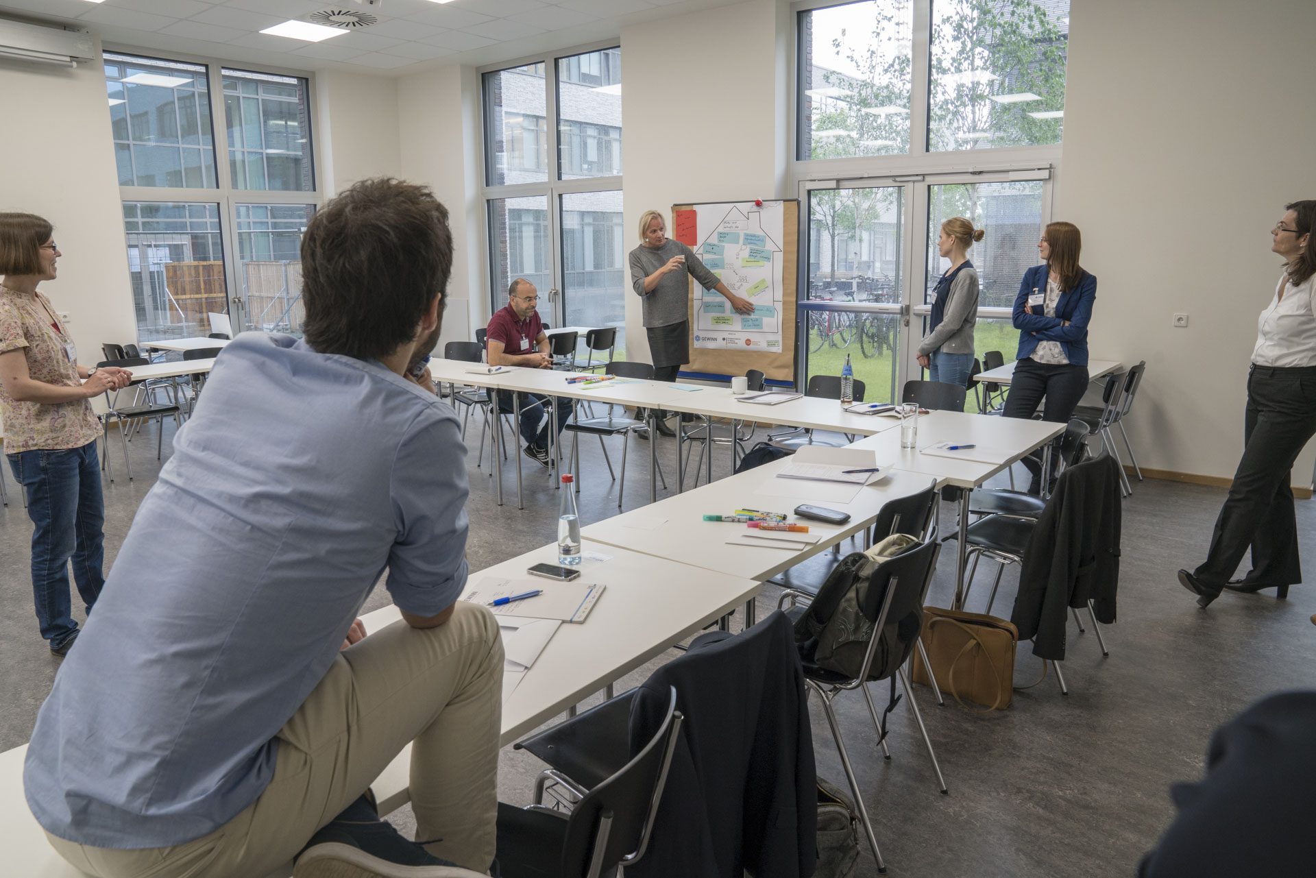 Gruppe von Personen verteilt im Raum lauschen Rednerin vor Stelltafel